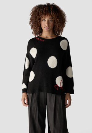 Frau mit lockigem Haar, die einen schwarzen Pullover mit großen weißen Punkten und roten bestickten Details trägt, kombiniert mit schwarzen Nadelstreifenhosen.