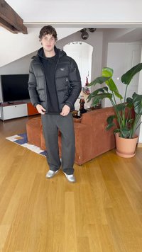 Casaco puffer preto com capuz e fechos, usado sobre um hoodie preto; calças cinzas e sapatilhas prateadas; ambiente interior com plantas.