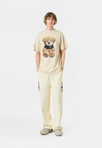 Camiseta beige de gran tamaño con un gráfico de un oso de peluche; pantalones color crema con acentos de oso; calzado cómodo; fondo neutro.