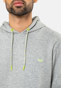 Grijze hoodie van zachte stof, met een trekkoorde met gele en grijze accenten en een klein groen logo op de borst.
