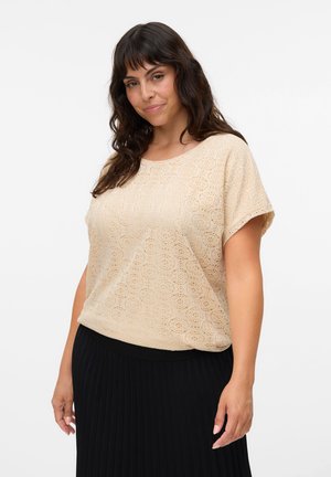 Femme aux cheveux bruns ondulés portant un haut beige en dentelle à manches courtes et une jupe noire plissée, debout devant un fond blanc uni.