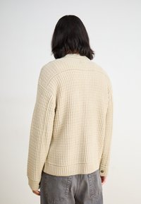 Beige adīta kardigāna ar strukturētu rakstu, nolaistām plecu daļām un ribotām piedurknēm. Aizmugures skats, izceļot auduma un konstrukcijas detaļas.