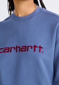 Sudadera azul claro con cuello redondo, con el logotipo "carhartt" bordado en color burdeos en el pecho sobre una tela suave.