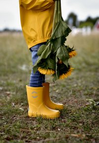 Gule gummistøvler med grå detaljer, båret over blå cowboybukser, stående på græs og holdende solsikker med grønne blade.