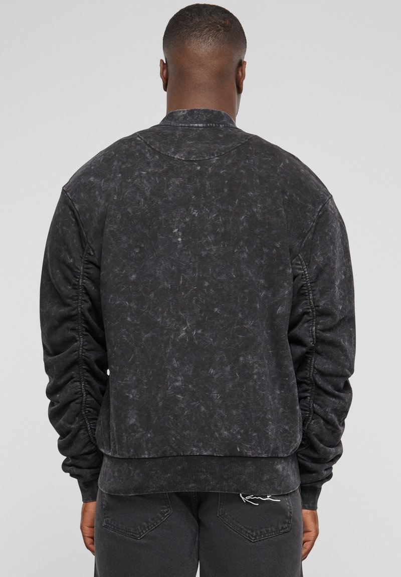 Giacca bomber nera, con texture e finitura sfumata, polsini e orlo a coste. Presenta un collo arrotondato e dettagli elasticizzati sulle maniche.
