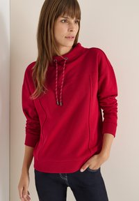 Czerwony hoodie z wysokim kołnierzem, z akcentami w postaci sznurka i ściągaczy na rękawach. Gładka tkanina z subtelnymi szwami, noszona na ciemnych dżinsach.