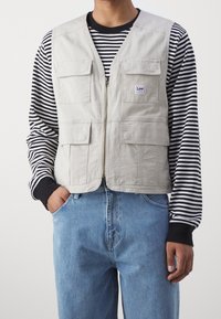 Gilet grigio chiaro con molte tasche frontali e chiusura con zip, indossato sopra una camicia a righe bianco e nere. Jeans in denim blu visibili sotto.
