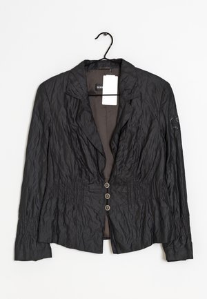 Blazer en tissu noir froissé avec trois boutons, col à revers cranté, manches longues et un détail brodé sur la manche gauche, suspendu à un cintre.