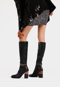 Stivaletti neri in pelle al ginocchio con tacco a blocco, caratterizzati da un accentuato in catena dorata e una fascia marrone sul tallone per contrasto. Texture liscia.