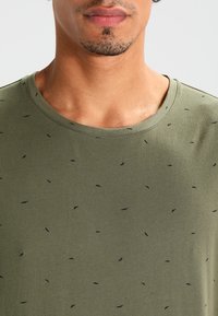 Olivengrøn t-shirt med rund halsudskæring og små sorte bladlignende mønstre. Lavet af blødt stof med en glat tekstur.