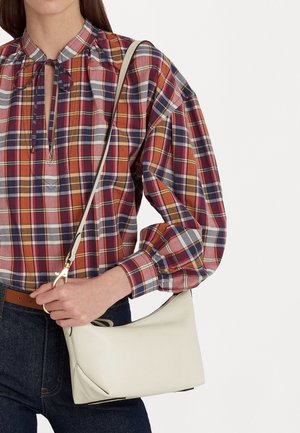 Femme portant un haut à carreaux rouge, bleu et jaune avec des manches bouffantes, tenant un petit sac à épaule blanc et portant un jean foncé avec une ceinture marron.