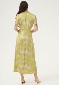 Maxi šaty s světle béžovým pozadím a velkým vzorem zeleného monstera listu. Kapsové rukávy a zapínání na knoflík na krku.
