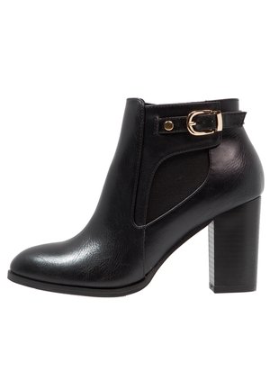 Bottine en cuir noir avec talon bloc, panneau élastique latéral et détail de sangle à boucle dorée près du haut.