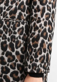 Acercamiento a la manga de un abrigo de lana con estampado de leopardo, con un bolsillo de ribete y la muñeca de una persona visible en el puño.