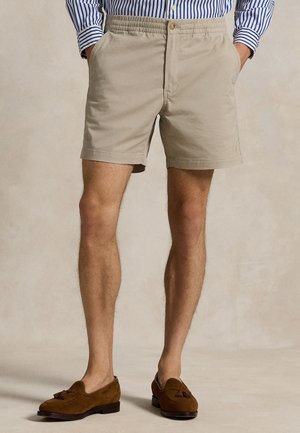 Man draagt beige shorts met elastische taille, een blauw-wit gestreept overhemd in de broek gestopt en bruine suède loafers met kwastjes, handen in de zakken.