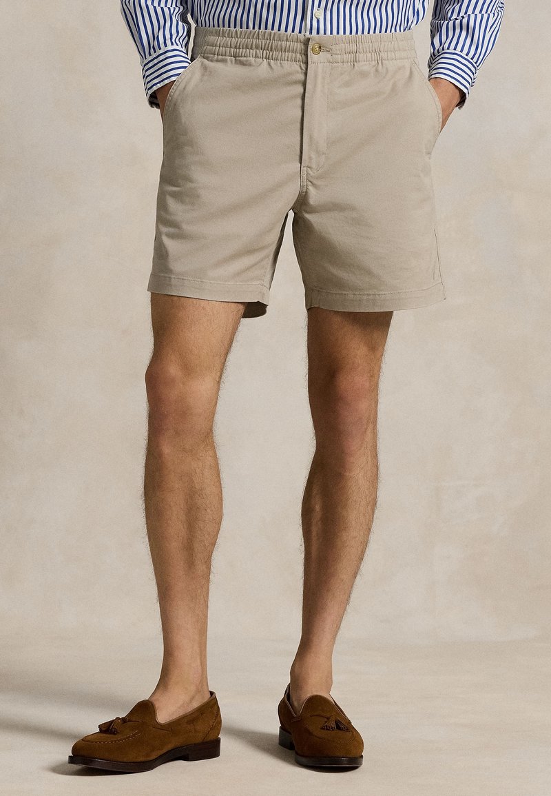 Homme portant un short beige à taille élastique, une chemise rayée bleue et blanche rentrée, et des mocassins en daim marron à pampilles, mains dans les poches.