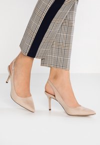 Escarpins beige à talons ouverts avec un bout pointu, en tissu lisse, et un fin talon aiguille, associés à un pantalon à carreaux bien ajusté.