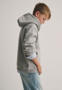 Grauer Kapuzenpullover mit strukturiertem Stoff, gerippten Bündchen und Seitentaschen. Darunter ist ein blau-weiß gestreiftes Hemd sichtbar.