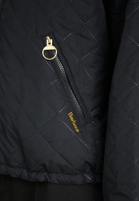 Ruutudega must jakk diagonaalsete õmblustega, millel on kuldne tõmblukk ja kollane "Barbour" logo ääres. Pehme tekstuur, kerge disain.
