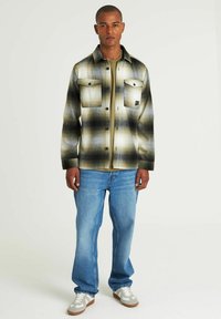 Camicia flanella a quadri in grigio e nero, con taschini sul petto e bottoni, indossata sopra una camicia verde, abbinata a jeans blu e sneakers grigie.