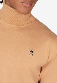 Beige coltrui van zacht breimateriaal. Bevat een ribgebreide kraag en een klein marineblauw logo geborduurd op de borst.
