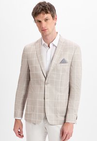 Mann in einem beigen Blazer mit Fensterrutenmuster, zwei Knöpfen, weißem Hemd, weißen Hosen und einem gemusterten Einstecktuch.