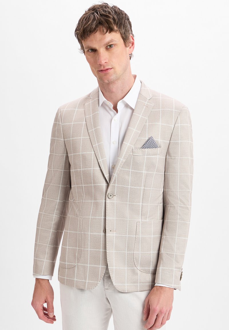 Mann in einem beigen Blazer mit Fensterrutenmuster, zwei Knöpfen, weißem Hemd, weißen Hosen und einem gemusterten Einstecktuch.