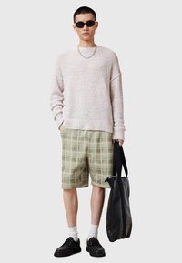 Sweater i lyserød tekstur, grønternet shorts, sorte sko og solbriller; holder en sort taske. Neutral baggrund.