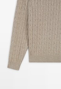 Světlý béžový svetr s copánkovým vzorem, žebrovanými manžetami a lemem. Vyrobený z měkké látky, s volnějším střihem a texturovaným povrchem.