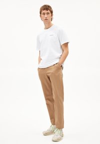 T-shirt en coton blanc avec un petit logo bleu, associé à un pantalon marron clair et des baskets à motifs avec des accents verts.