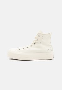 Λευκά καμβά αθλητικά παπούτσια υψηλής κορυφής με υφή στο καουτσούκ σόλες, μεταλλικές οπές και διακοσμητικά τρουκς στο πλάι. Ισιωμένα κορδόνια και στρογγυλή μύτη.