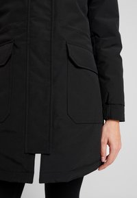 Černá parka s dvěma velkými předními kapsami, zaobleným lemem, elastickými manžetami a hladkým matným povrchem. Obsahuje zip a knoflíkovou légu.