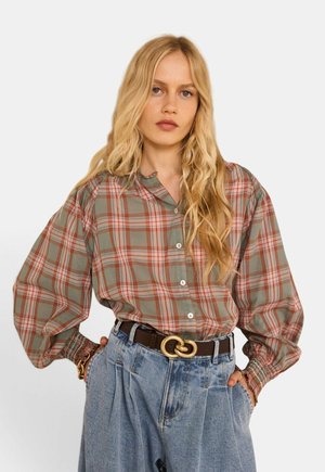 Femme blonde portant une chemise à manches longues ample à carreaux, rentrée dans un jean bleu taille haute avec une ceinture noire et une boucle dorée.