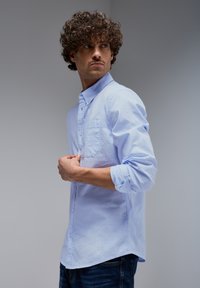 Light blue, long-sleeve button-up shirt with a chest pocket. Smooth cotton fabric, relaxed fit, sleeves rolled up to the elbows, paired with dark jeans.