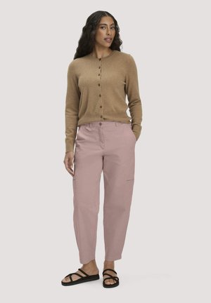 Frau mit langen lockigen Haaren, die eine braune, geknöpfte Strickjacke, hellrosa Hosen und schwarze, Riemchensandalen trägt, steht mit den Händen in den Taschen.