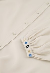 Crème kleurige stof met een rij knopen, voorzien van een geplooide mouwmanchet versierd met kleurrijke sterrenborduursels in blauw, zwart en goud.
