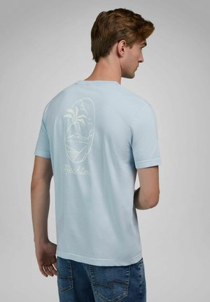 Junger Mann von hinten gesehen, trägt ein hellblaues T-Shirt mit einem Palmen- und Surfbrettmotiv über dem Schriftzug "Hechter" und eine blaue Jeans.