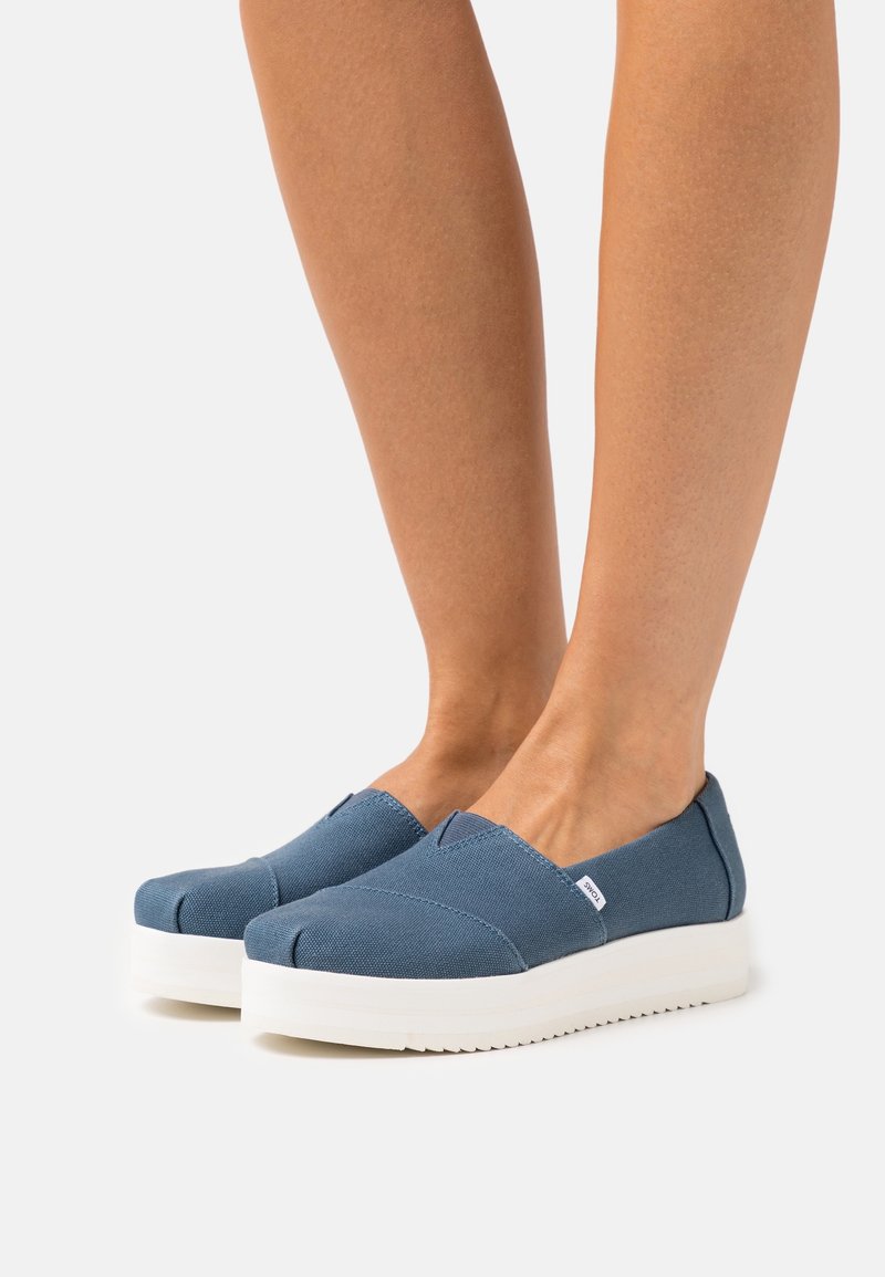 Jambes portant des chaussures en toile bleues à enfiler avec des semelles épaisses en caoutchouc blanc sur fond blanc.