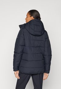 Marineblå polstret jakke med hette, med horisontalt quiltet design, glatt tekstur og elastiske mansjetter. Ingen synlige maskinvare detaljer.
