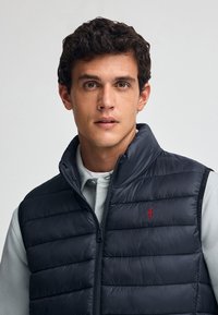 Granatowa kamizelka puchowa z poziomymi przeszyciami, z przodu na zamek błyskawiczny oraz czerwoną logo na piersi. Założona na jasnoszary, długi rękaw.
