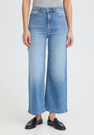 Mujer que lleva vaqueros de pierna ancha azul claro con cintura alta, botón y cremallera delanteros, combinados con mocasines negros y anillos en los dedos.