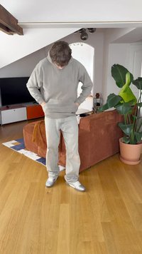 Felpa grigia, jeans di denim chiaro e sneaker argentate. La scena include un divano marrone a coste, una pianta da interno e un pavimento in legno.