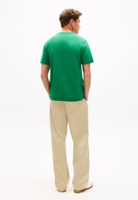 Žalia medvilninė marškinėliai su apvaliu kaklu, trumpomis rankovėmis ir subtiliu nugaros detalumu, derinami su smėlio spalvos plačiomis kelnėmis.
