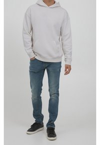 Sweat à capuche blanc avec une poche avant, associé à un jean bleu clair et des baskets noires. Design simple sans motifs ni accents visibles.