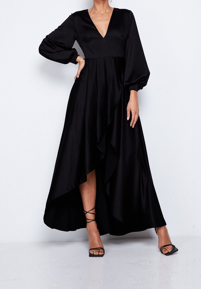 Kvinna i en svart långärmad, flödande klänning med en djup V-ringning och asymmetrisk fåll, kombinerad med svarta snörda högklackade sandaler.