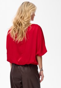 Blonde vrouw in een losse rode blouse en donkere leren broek, gezien van achteren, met een zichtbare tatoeage op haar rechter onderarm.