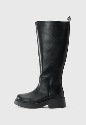 Aukštas juodas odinis botas su kilpike viršuje, apvaliu pirštu ir storu žemu kulnu priešais paprastą baltą foną.