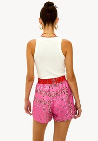 Top sin mangas blanco combinado con pantalones cortos estilo bandana en rosa que presentan patrones de paisley y una banda elástica roja en la cintura. Modelo de espaldas.