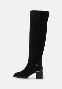 Botas negras de ante por encima de la rodilla. Punta redonda, tacón grueso y acento de logotipo dorado en el costado. Textura suave con un diseño ajustado.