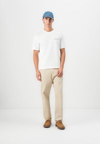 White cotton t-shirt with logo, beige trousers, and brown suede shoes. Light blue cap, standing against a plain white background.
